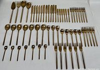 Full layout of entire 56 piece bronze flatware set including knives, forks, spoons, salad servers and teaspoons on white background.