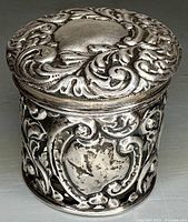 Front and side view of the ornate sterling silver canister showing repoussé floral and scroll designs and shield cartouche on the side.