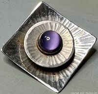 Close-up of front of square silver-tone brooch with central purple cabochon stone showing textured hammered metal.