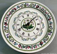 Front view of round ceramic wall clock showing decorative floral border and Roman numeral clock face with black hands and floral center.