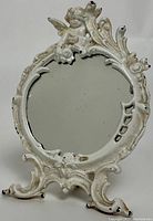 Front view of the oval standing mirror showing the white painted cast-iron frame with cherub figure on top and ornate Rococo-style detailing.