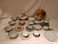 Full lot overview showing the assortment of 19 Asian themed cups, saucers, bowls, and plates arranged on a white cloth background.