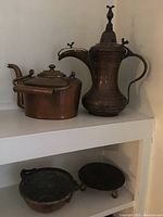 Photo of copper decorative lot items on white shelf including two teapots, small hammered pot and small pan, shown with and without soda can for scale.