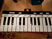 Top-down view of the floor piano mat showing 37 keys with illustrated instruments above keys and buttons for volume control, record, demo, playback, and play functions.