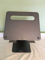 Front view of matte black metal laptop stand showing adjustable height and ventilation slots, with rubber pads on corners.