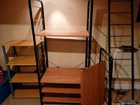 Photo showing two shelving units side by side. Smaller unit has 3 wooden shelves with silver legs, larger unit has black metal frame with wooden shelves and extra shelves leaning against it.