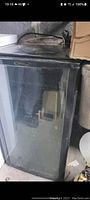 Black compact wine fridge with transparent glass front door, showing surface wear and scuffs