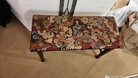 Top view of the piano bench seat showing intricate needlepoint upholstery with colorful and detailed design including a figure and floral patterns.