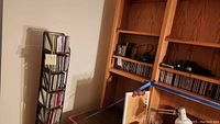 Photo showing a tall metal CD rack filled with CDs, adjacent to a wooden shelving unit with more CDs and electronic items