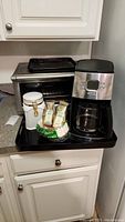 Photo showing Beeville toaster oven with tray on top and Hamilton Beach coffee maker on black tray with coffee accessories including packets, filters, and small jar.