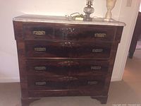 Front view of dark wood four drawer dresser with marble top, brass decorative drawer pulls, and carved drawer motifs