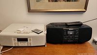 Side-by-side photo of white Henry Kloss Model 88CD radio and CD player and black Sony CD/radio/cassette corder with their remotes on a wooden surface.
