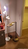 Photo showing two standing pole lamps; one brass Verilux lamp with flat tray and adjustable arm, the other white lamp with multiple lights.