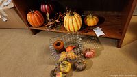 Photo showing multiple artificial pumpkins of varying sizes on a wooden shelf, several gourds scattered on carpet, and a rectangular clear glass container with circular bubble patterns containing a large brown scented candle and some gourds.