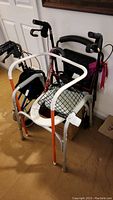 Photo showing two walkers - a 3-wheeled Nova walker with orange frame and mesh seat and a black Drive rollator with four wheels and seat, also visible two canes and part of the grabber and bed assist rail behind.