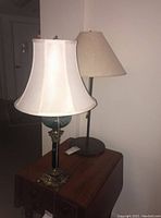Photo of two lamps side by side on a wooden table against a wall. One lamp has a green textured glass globe on the stem, white fabric bell-shaped shade, and ornate brass base. The other lamp has a plain beige conical shade and simple dark stem and base.