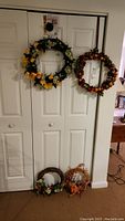 Photo showing two large wreaths hanging on closet doors and two smaller wreaths on the floor beneath, featuring autumn and floral designs.