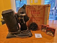 Photo of the Excel 16mm movie projector on a wooden table with its original box in the background, a metal film reel and two sealed film boxes beside it. Power cord is wrapped on projector base. The box has printed text 'MODEL EXCEL 16 M.M. MOTION PICTURE PROJECTOR'.