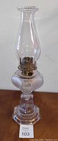 Full view of the antique glass oil lamp standing upright showing detailed stem, base, and tall chimney.