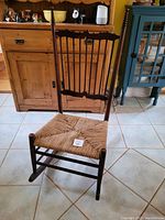 Front angled view of rocking chair showing woven rope seat and ornate finials on back posts.