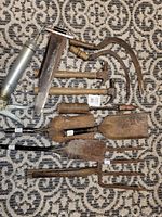 Photo showing assorted old tools on patterned carpet, including metal shovels, hammers, soldering tool, scythe, and smaller hand tools, some with wooden handles