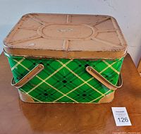 Closed view of rectangular green plaid metal lunch basket with dual metal handles resting on wooden surface.
