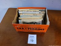 View of the over 200 vintage postcards stored upright in an orange Lea & Perrins Worcestershire Sauce cardboard box on a wooden table, showing the quantity and storage method.