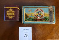 Top-down view of two antique smoker's tins: John Middleton's Club Mixture (square, maroon) and Player's Navy Cut Cigarettes (rectangular, blue with sailor artwork) with auction lot card