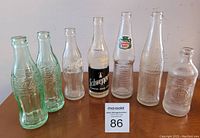 Photo of seven vintage glass soda bottles including Coca-Cola, Pepsi, Canada Dry, and Schweppes, arranged in a row on a wooden surface, all in good condition.