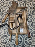 Photo of 8 old hand tools including hand drill, cleaver, rasp, long screwdriver, mallet, small plane, and two hammers arranged on carpet and in a small box