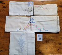 Photo of four folded antique white pillowcases with hand-stitched needlepoint designs arranged on wooden surface. Shows embroidery details such as a perched blue bird, multi-color flowers, and white-on-white floral needlework.