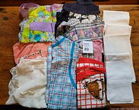 Photo showing seven aprons in various colors and designs, together with four white linen napkins folded and stacked on the right side on a wooden surface.