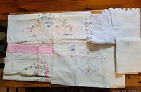 Nine vintage linen pillow cases displayed on wooden surface, showing various embroidered floral and needlepoint details, lace inserts, and different styles of edging and hems.