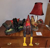 Photo showing all three rooster themed items on a wooden dresser with drawer handles: cast iron bookends, lamp, and wooden figurine with movable legs.