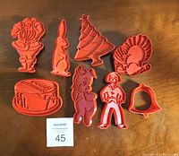 Photo showing eight red plastic cookie cutters laid out on a wooden surface including Santa Claus, Easter Bunny, Christmas Tree, Turkey, Happy Birthday cake, Pig, Human figure, and Bell shapes.