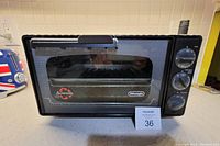 Black DeLonghi fan convection toaster oven with a mesh front glass door and control dials, shown closed on a kitchen counter.