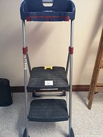 Front view of the Cosco two-step ladder showing black plastic steps, metal frame, and blue utility tray.