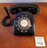 Front-facing photo of vintage black rotary dial telephone with coiled handset cord and visible phone number on dial center.