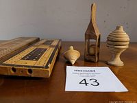 Photo showing two inlaid wooden cribbage boards, hand-carved ball puzzle in a narrow triangular frame, and two wooden spinning tops arranged on a wooden surface.