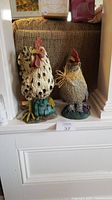 Front view of two rooster figurines on a ledge near window, one white with black spots, the other heavier with straw neck decoration.