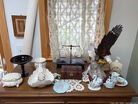 Full view of assortment on wooden surface with lace curtains background, showing eagle figurine, wooden chest, balance scale, various ceramics, and glass bowl.