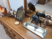 Wide view of dresser top showing an assortment of decorative items including a vintage ornate vanity mirror, bull figurines, a rectangular mirrored tray, small ornamental dishes and candle votive.