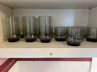 Thirteen smoke color beverage glasses arranged with taller glasses on the left and shorter on the right on a white shelf.