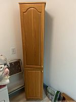 Front view of tall wood storage cabinet with two doors and gold-colored round knobs in a corner near a folded metal step stool.