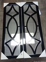 Front view of the two decorative wall mirrors, showcasing full reflective surface with black metal geometric overlay on a wood floor.