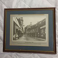 Framed black and white historical print titled 'White Hart Street, 1930' showing a street view from early 20th century with shops and signage. Frame is wood with gold inner trim, matting in blue and beige.