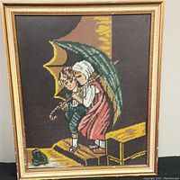 Framed needlepoint art showing two children sitting on wooden steps under an umbrella, with a visible gold frame and glass cover.