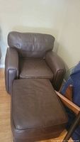 Brown leather chair with matching ottoman placed in room against pale wall, wooden floor visible.