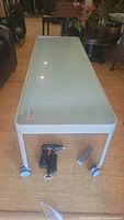Top view of rectangular media table with smoked glass surface and white metal frame on four locking caster wheels. Light reflects on glass.