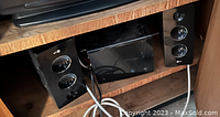 Photo showing two black LG speakers and one LG black subwoofer placed inside a wooden shelving unit.